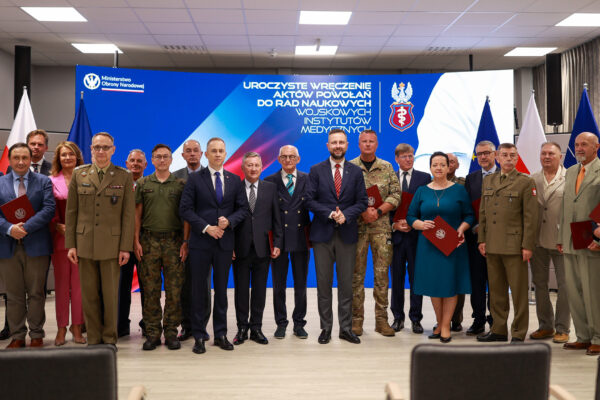 Profesor Waldemar Machała, w ciemnym garniturze z niebieskim krawatem, stoi wśród kilkunastu osób – wojskowych w mundurach i cywilów – podczas uroczystego wręczenia aktów powołania do Rad Naukowych Wojskowych Instytutów Medycznych. W tle duży ekran z logotypem Ministerstwa Obrony Narodowej i informacją o wydarzeniu.