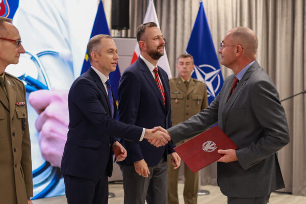 Profesor Waldemar Machała, ubrany w szary garnitur i czerwony krawat, odbiera akt powołania od sekretarza stanu w MON, Cezarego Tomczyka, w obecności ministra Władysława Kosiniaka-Kamysza. W tle flagi Polski, NATO i Ministerstwa Obrony Narodowej.