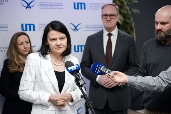 Kobieta stoi przy mikrofonie udzielając wywiadu, dwie osoby w tle