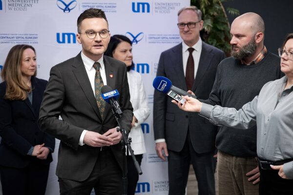 Mężczyzna stoi przy mikrofonie udzielając wywiadu, trzy osoby stoją za nim