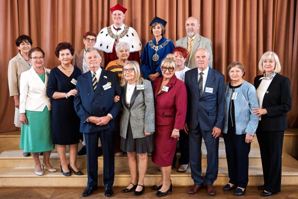 Mniejsza grupa kilkunastu absolwentów ustawiona na schodach w eleganckiej sali. Wśród uczestników znajdują się także osoby w stroju akademickim z łańcuchami, pozostali uczestnicy ubrani są formalnie.