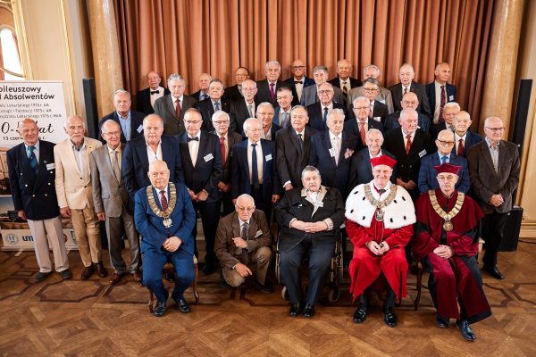 Duża grupa absolwentów w formalnych strojach ustawiona do zdjęcia w kilku rzędach Po prawej stronie widoczne osoby w czerwonych togach z łańcuchami akademickimi, a po lewej baner informujący o jubileuszowym zjeździe absolwentów.