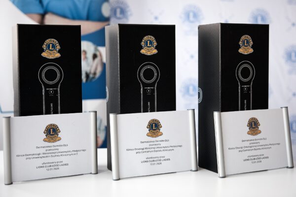 Trzy czarne pudełka z dermatoskopami stoją obok siebie. Przed nimi znajdują się tabliczki z informacją o darowiźnie dla klinik Uniwersytetu Medycznego w Łodzi.