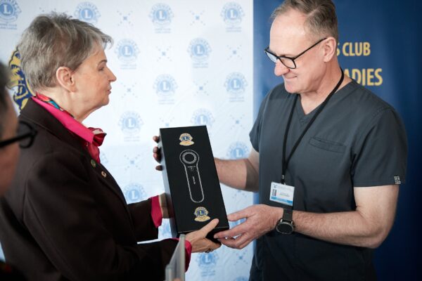 Osoba w stroju formalnym przekazuje czarne pudełko z dermatoskopem osobie w ubraniu medycznym. W tle widoczny baner z logo „Lions Club Łódź-Ladies”.