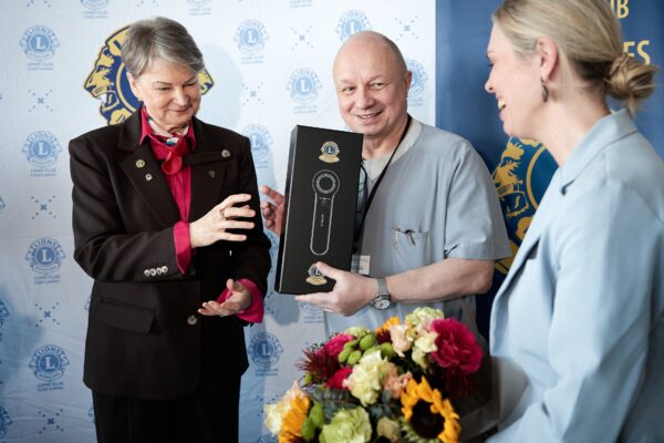 Osoba w stroju formalnym rozmawia z osobą w ubraniu medycznym, która trzyma pudełko z dermatoskopem. Obok stoi kolejna osoba z bukietem kwiatów. W tle baner z logo „Lions Club Łódź-Ladies”.