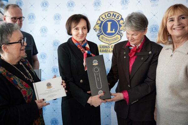Osoba w stroju formalnym przekazuje pudełko z dermatoskopem innej osobie. Obok widoczna jest tabliczka z informacją o darowiźnie. W tle baner „Lions Club Łódź-Ladies”.