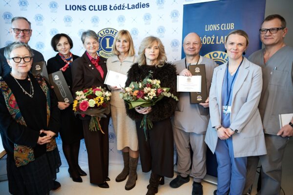 Grupa osób stoi na tle baneru z logo „Lions Club Łódź-Ladies”. Kilka osób trzyma czarne pudełka z dermatoskopami oraz tabliczki z informacją o darowiźnie. Widać także dwa bukiety kwiatów.