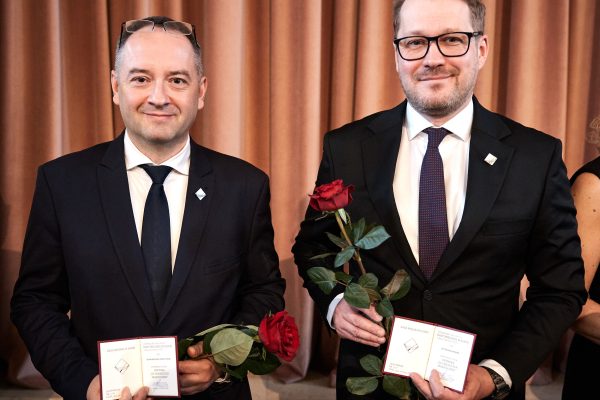 Dwóch mężczyzn w garniturach trzyma w ręku odznakę, legitymację i czerwoną różę