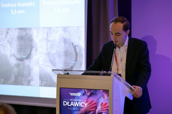 Mężczyzna w garniturze przemawia przy mównicy podczas konferencji „Akademia Dławicy”. Za nim widoczny jest slajd z obrazami angiograficznymi i informacją o średnicy naczynia